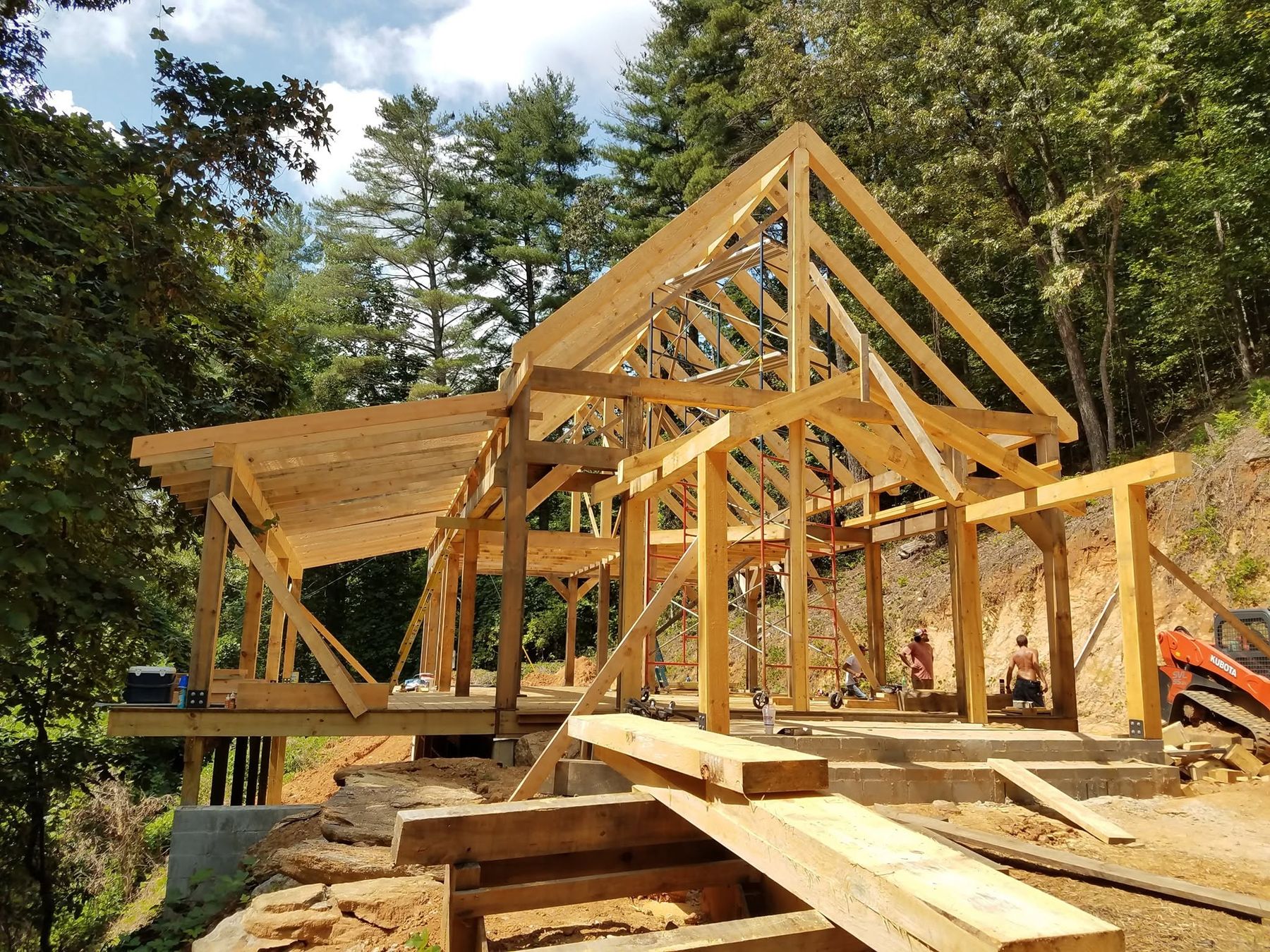 A timber frame being raised on a wooded hillside, with fresh-cut beams stacked in front