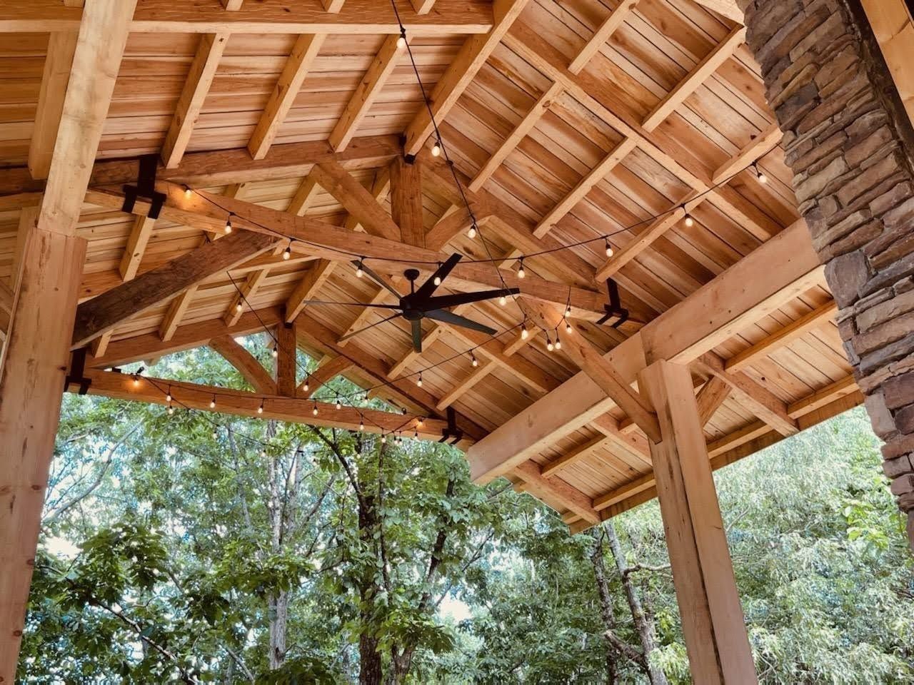 Looking up into a timber pavilion roof: heavy posts, braces and trusses under a plank ceiling, with string lights and trees beyond