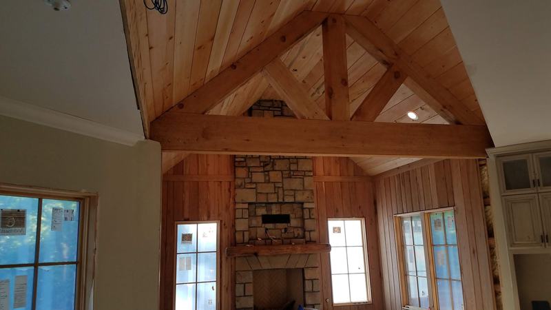 A timber truss and wood plank ceiling over a stone fireplace with a wood mantel. Opens larger.