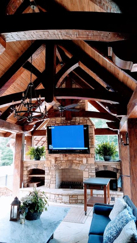A covered outdoor room with dark timber trusses over a stone fireplace. Opens larger.